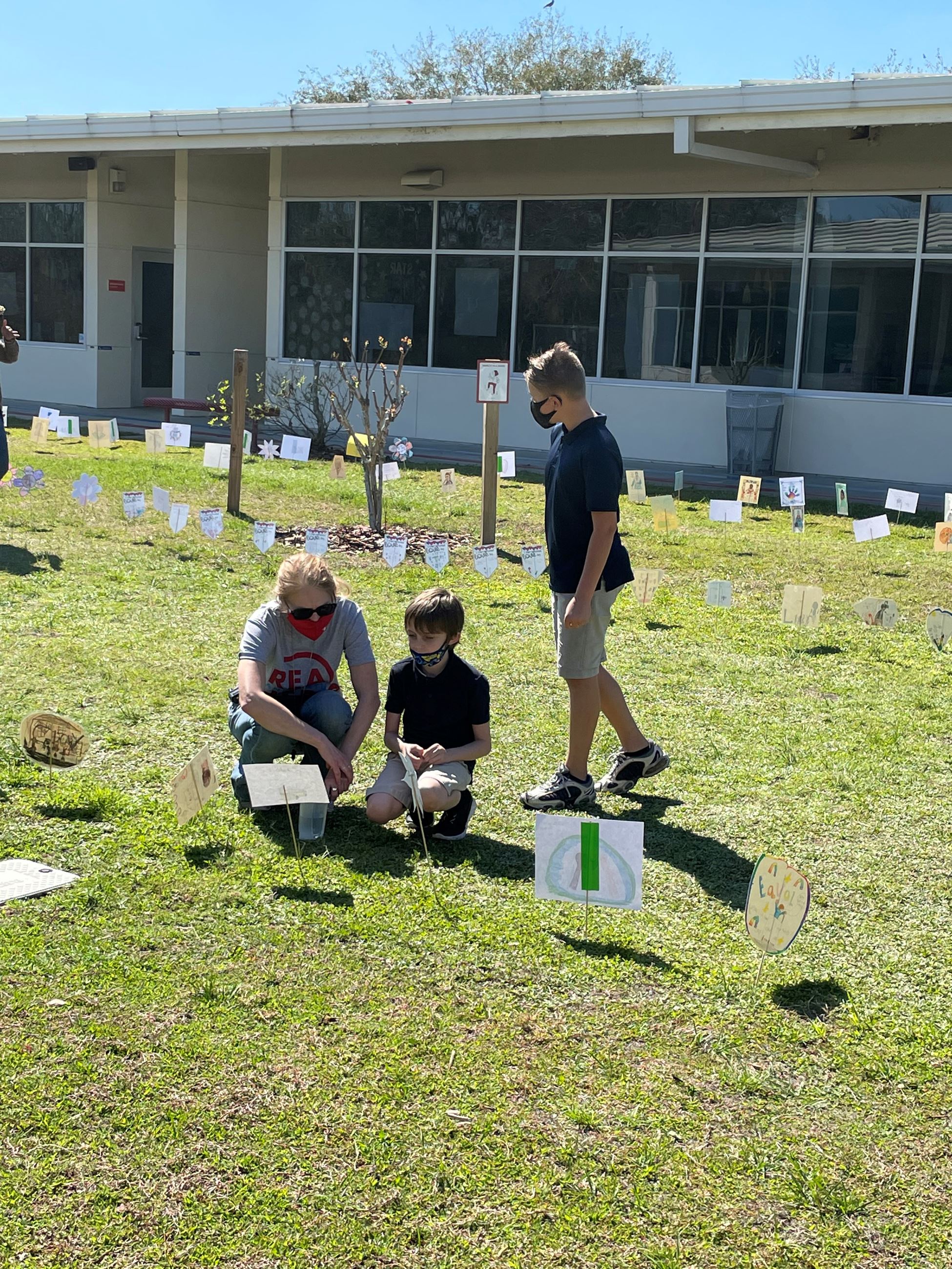 A female teacher kneeling with a young boy admiring student artwork outside in the courtyard. 