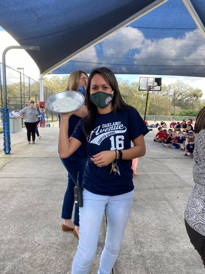 A female teacher wearing an OACS tee shirt holding a pie ready to throw it at somone!